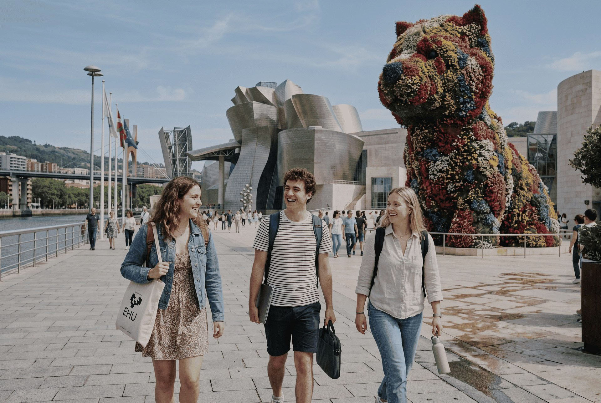 Tres estudiantes internacionales caminan sonrientes por el paseo junto al Museo Guggenheim de Bilbao en un día soleado. Al fondo se distingue la icónica arquitectura del museo y la escultura floral Puppy, mientras los estudiantes llevan mochilas y material de estudio, reflejando una experiencia académica y cultural en la ciudad.